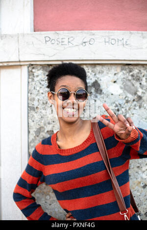 Portrait von lächelnden jungen Frau eine Sonnenbrille trug und gestreifter Pullover mit Sieg Zeichen Stockfoto