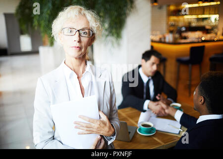 Portrait von Reife Geschäftsfrau in Brillen Holding business Vertrag mit ihren Partnern die Hände schütteln im Hintergrund Stockfoto
