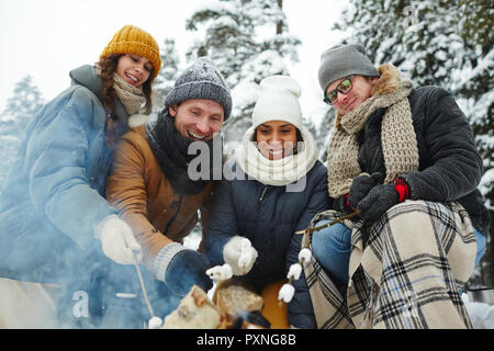 Positiv begeistert Junge multi-ethnischen Freunde im Winter Mäntel Sitzen am Lagerfeuer Marshmallows rösten und auf Feuer auf dem Campingplatz im Wald Stockfoto