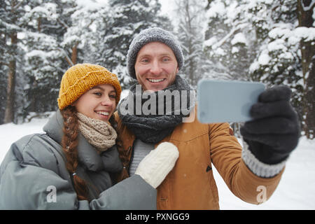 Glücklich, aufgeregt, junges Paar in warmen Hüte verinnerlichen und sich Fotografieren auf dem Smartphone beim Gehen in Winter Park Stockfoto