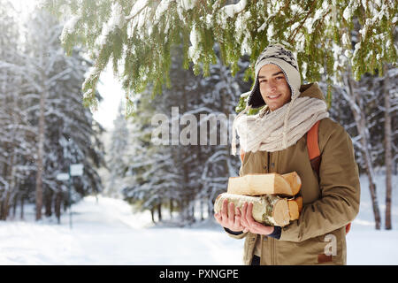 Taille bis Portrait von gutaussehenden jungen Mann im Winter Wald holding Protokolle aufwirft und an der Kamera freundlich lächelnd, Kopie Raum Stockfoto
