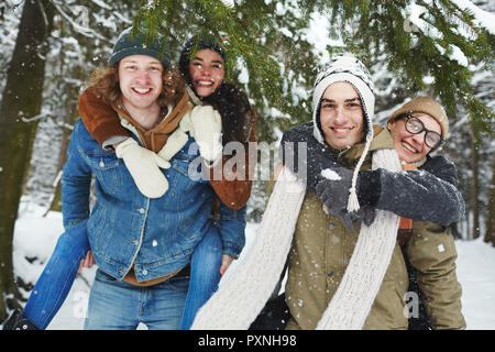 Porträt von zwei glückliche junge Paare Spaß auf Winter Resort und in die Kamera lächeln, während piggyback spielen in den verschneiten Wald Stockfoto