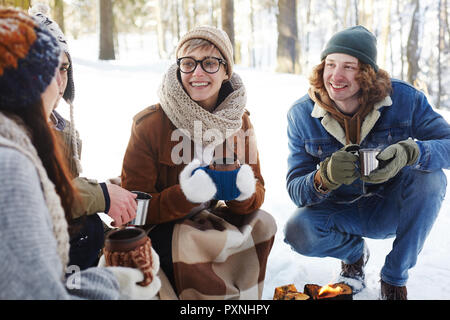 Portrait von glücklichen jungen Menschen Camping im Winter Wald im Kreis um das Feuer sitzen und plaudern, während Sie cups Stockfoto