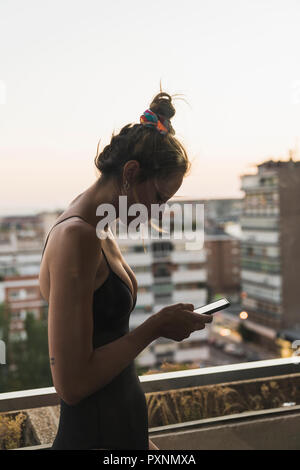Junge Frau mit schwarzen Kleid mit Handy auf dem Balkon Stockfoto