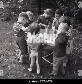 1950er Jahre, junge Kinder in einer spielgruppe oder Kindergarten draußen Spaß zusammen hat das Spielen mit alten Spülmittel, Flaschen und Wasser in einer großen Schüssel, eine Aktivität, die Entdeckung und sensorischen Fähigkeiten Entwicklung. Stockfoto