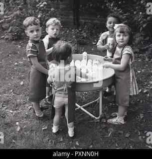1950er Jahre, junge Kinder in einer spielgruppe oder Kindergarten draußen Spaß zusammen hat das Spielen mit alten Spülmittel, Flaschen und Wasser in einer großen Schüssel, eine Aktivität, die Entdeckung und sensorischen Fähigkeiten Entwicklung. Stockfoto