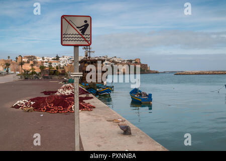 Cal Wasser der Bucht mit geparkten Boote. Bürgersteig mit einem Zeichen der verbotenen Angeln. Stapel der Fischernetze. Die Gebäude der Stadt im Hintergrund. Bewölkt Stockfoto