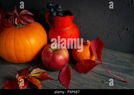 Zwei Kürbisse mit hellen Blätter im Herbst Apfel, Trauben und roten Kanne auf Holz- rustikalen Tisch gegen einen schwarzen rauen Beton grunge Wand. Herbst Ernte und Stockfoto