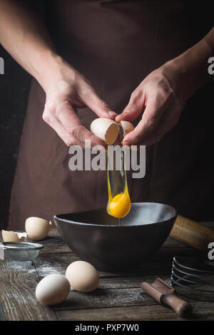 Professioneller Koch Hände brechen ein Ei in die Schüssel Teig auf hölzernen Tisch zu machen, über die dunklen Hintergrund Stockfoto