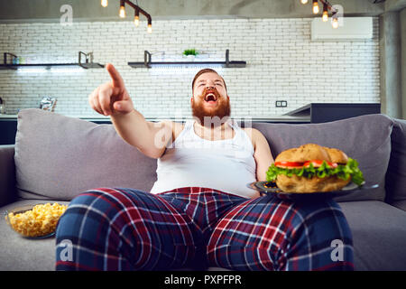 Dicke lustiger Mann mit einem Burger sitzen auf der Couch. Stockfoto
