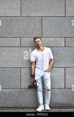 Bild von Stattlichen kaukasischen Kerl auf Grau texturierte Wand im weißen T-Shirt und eine weisse Jeans mit Kamera am Gurt auf seinem Arm. Stockfoto