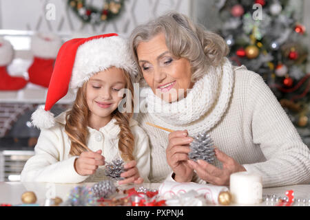 Portrait von Happy Großmutter und ihre kleine Enkelin Vorbereitung auf das Weihnachtsfest gemeinsam zu Hause, malen Tannenzapfen Stockfoto