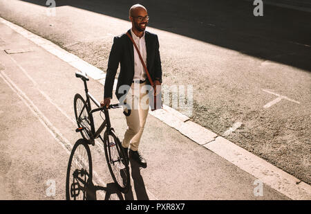 Hübscher junger erwachsener Mann mit Anzug zu Fuß mit seinem Fahrrad im Freien. Afrikanischen Geschäftsmann, Büro mit seinem Fahrrad. Stockfoto