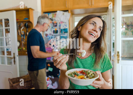 Gerne reife Frau zu Hause essen einen Salat mit Mann im Hintergrund Stockfoto