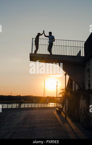 Zwei Freunde, high-fiving bei Sonnenuntergang, stehend auf der Aussichtsplattform Stockfoto