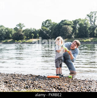 Vater und Sohn Spaß am Flußufer, spielen mit einer Wasserpistole Stockfoto