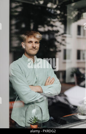 Junge Mann am Fenster, mit verschränkten Armen, mit Ohrstöpsel Stockfoto