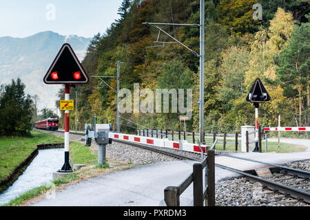 Railraod Kreuzung mit blinkenden Warnlampen und einem roten Zug nähert sich in der Schweizer Landschaft Stockfoto