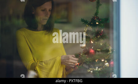 Junge Frau im gelben Hemd schmücken Weihnachtsbaum im Innenbereich. Blick durch ein Fenster. Stockfoto