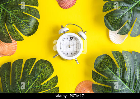 Kleine Wecker mit tropischen Palmen und Muscheln auf gelben Hintergrund. Sommer minimal kreatives Konzept. Stockfoto