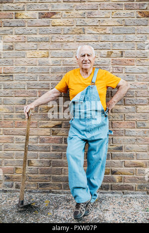 Älterer Mann in Jeans Overall mit Garten Werkzeug suchen Bei camera lehnte sich an eine Wand Stockfoto