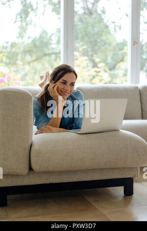 Lächelnd, reife Frau zu Hause auf der Couch liegend mit Laptop Stockfoto