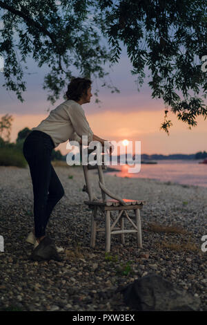 Frau lehnte sich auf einen Stuhl am Fluss, Sonnenuntergang beobachten Stockfoto