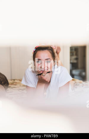 Portrait von lächelnden jungen Frau auf dem Bett zu Hause holding Pen liegen Stockfoto