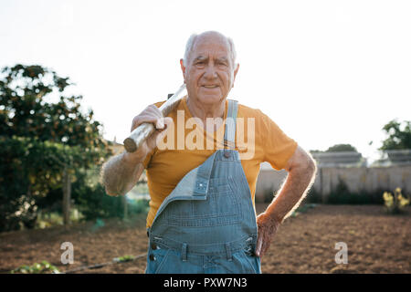 Älterer Mann in Jeans overall Holding Garten Werkzeug auf der Schulter und an der Kamera Stockfoto