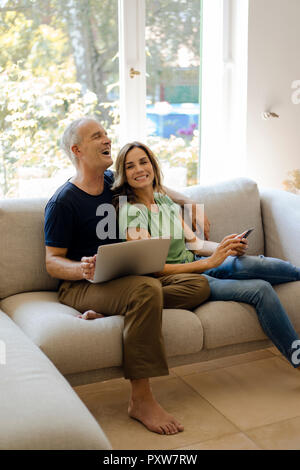 Glückliches junges Paar auf der Couch zu Hause mit Handy und Laptop Stockfoto