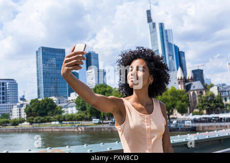 Deutschland, Frankfurt, Portrait von Inhalt junge Frau mit lockigem Haar unter selfie vor der Skyline Stockfoto