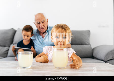 Portrait von kleinen Mädchen mit einem Glas Limonade zu Hause mit Bruder und Großvater im Hintergrund Stockfoto