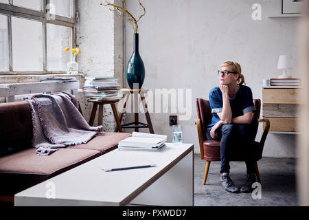 Nachdenkliche Frau sitzt auf einem Sessel im Loft auf der Suche durch das Fenster Stockfoto