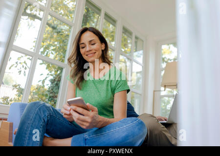 Smilong reife Frau auf der Couch zu Hause sitzen mit Handy mit Mann im Hintergrund Stockfoto