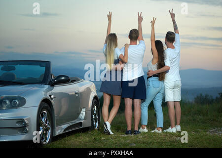 Zurück Blick auf zwei Paare stehen bewundern, tolle Aussicht und Landschaften, holding hands up und Frieden. Mann die Arme um Mädchen Taillen. Freunde in der Nähe von Silver cabriolet posieren. Stockfoto