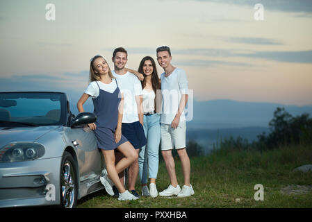 Unternehmen aus zwei Mädchen und zwei Männer stehen, Posing, Kamera und lächelnd. Schöne Mädchen in gestreiften Kleid und weißes T-Shirt lehnte sich auf Silber Cabrio. Freunde genießen Ausflug mit dem Auto. Stockfoto