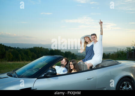 Gruppe von zwei Paare gehen für Laufwerk auf luxuriösen Silber Cabrio. Schöne Mädchen in weißen Rand breite Hut sitzen mit Jungen auf Fahrzeug, in die Kamera schauen und lächeln. Stockfoto