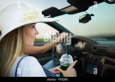 Seitenansicht der charmante junge Frau im weißen Rand breite Hut mit Blume sitzt im Cabrio. Adorable blonde Modell weg schauen und halten Limonade Glas. Mädchen sitzt in der Nähe der Fahrer. Stockfoto