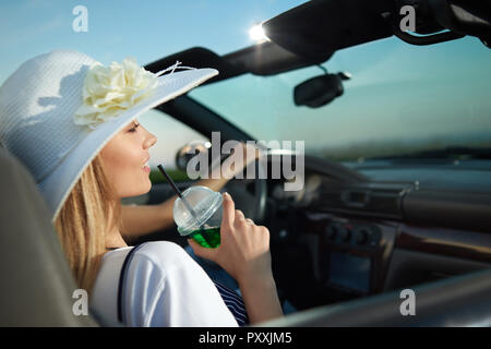 Wunderschöne Blondine Modell sitzen im Cabrio in der Nähe von Fahrer und weg schauen. Charmante junge Frau mit Limonade Glas und lächelnd. Mädchen tragen weiße Krempe breiten Hut mit Blume. Stockfoto