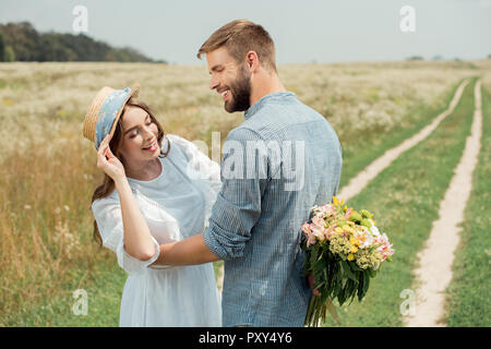 Lächelnd Mann verstecken Bouquet von wilden Blumen für Freundin hinter Zurück im Sommer Feld Stockfoto