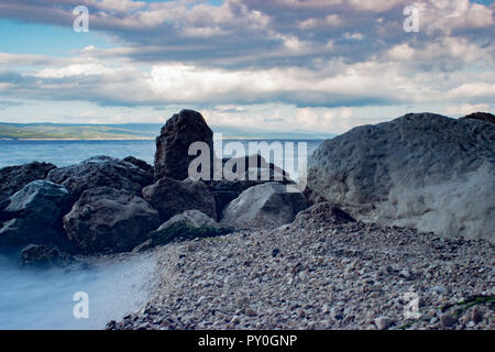 Surf Wellen des Meeres in Kroatien mit Steinen am Strand. Wellen gegen die Felsen und gesprüht. Im Hintergrund sind dunkle Wolken am blauen Himmel. Stockfoto