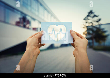 Nahaufnahme von Frau Hände halten ein Blatt Papier mit dem Herz Symbol innen, über Krankenhaus Gebäude Hintergrund. Gesundheitswesen und Medizin Konzept Stockfoto