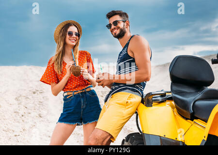 Glückliches junges Paar mit ATV und Kokosnuss Cocktails in der Wüste Stockfoto