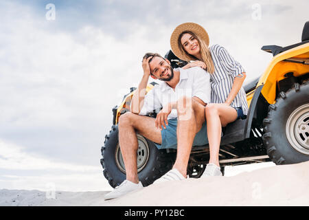 Schöne junge Paare sitzen auf ATV auf sandigen Dünen und Kamera Stockfoto