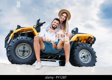 Ansicht von unten auf das glückliche junge Paare sitzen auf ATV auf sandigen Dünen vor Wolkenhimmel Stockfoto