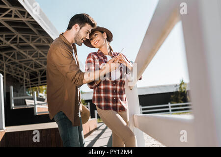 Lächelnd stilvolle Cowboy und Cowgirl in Freizeitkleidung auf Smartphone an der Ranch suchen Stockfoto