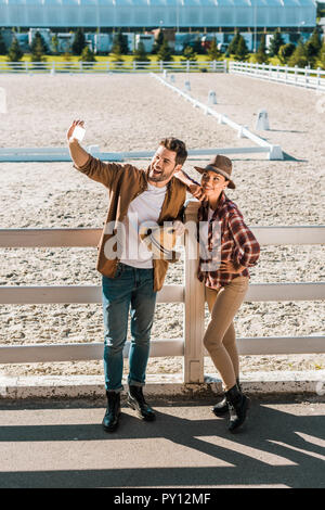 Lächelnd stilvolle Cowboy und Cowgirl in Freizeitkleidung unter selfie mit Smartphone an der Ranch Stockfoto