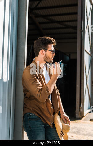 Seitenansicht des gutaussehenden Mann in Sonnenbrille rauchen Zigarette und lehnte sich an die Wand an der Ranch Stockfoto