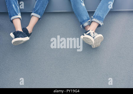 Teilweise mit Blick auf die paar Beine in Jeans und Turnschuhe sitzen auf graue Wand Stockfoto