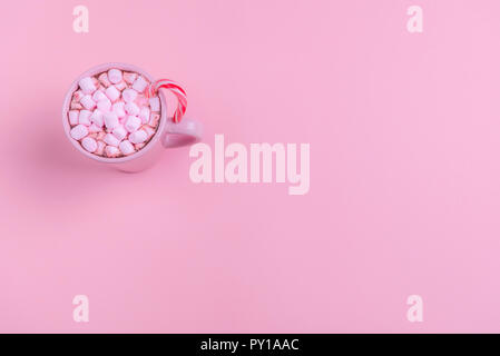 Weihnachten Kontext in einem minimalistischen Stil mit einem rosa Tasse heiße Schokolade, mini Marshmallows und ein Weihnachten Zuckerstange, auf rosa Papier Hintergrund. Stockfoto